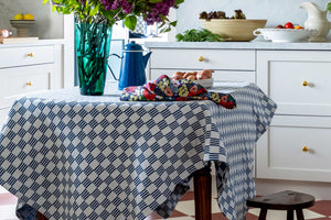 a peek into a cottagecore kitchen with a rustic table set with a blue and white checked tablecloth and lilacs, onions and other ingredients charmingly placed.