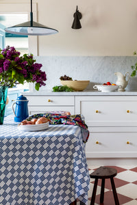 a peek into a cottagecore kitchen with a rustic table set with a blue and white checked tablecloth and lilacs, onions and other ingredients charmingly placed.
